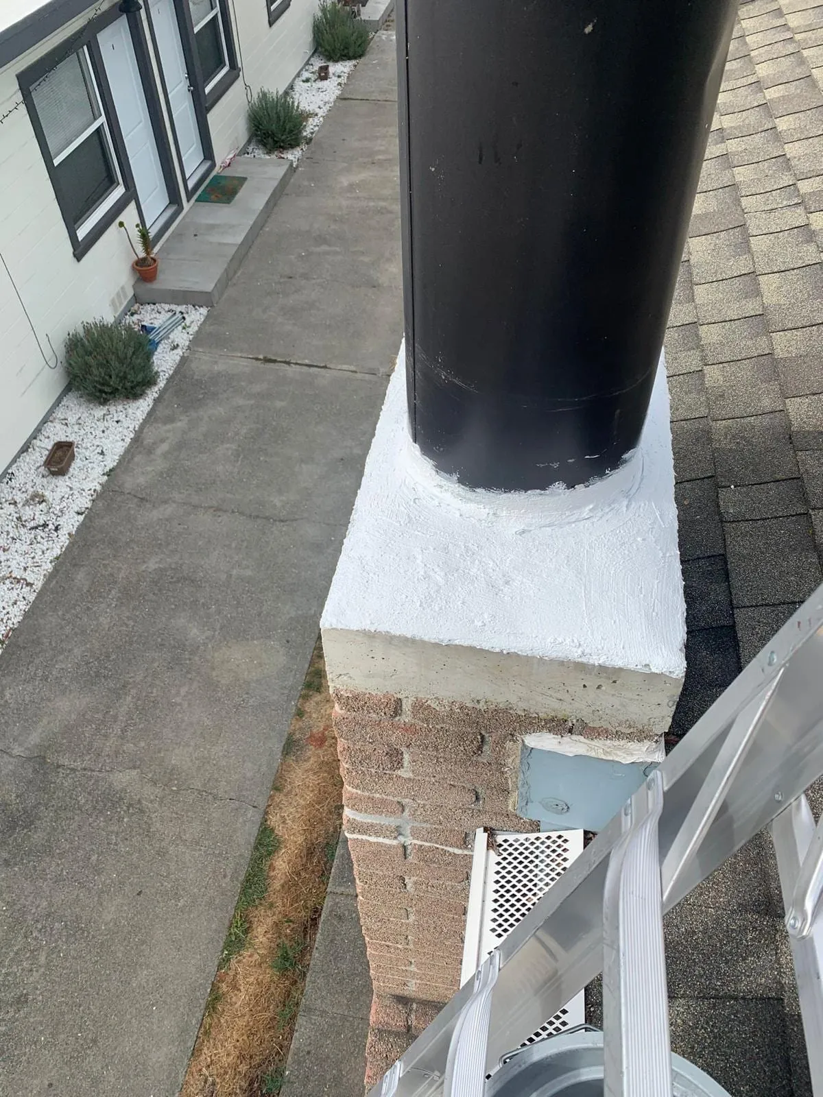 A black metal flue pipe rising from a brick chimney whose crown is coated in white sealant, with an aluminum ladder at the lower right